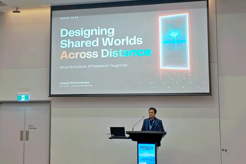 Dr. Yasas Sri Wickramasinghe smiling at the NZGDC 2026 lectern beneath his title slide, ‘Designing Shared Worlds Across Distance — What Multiplayer AR Research Taught Me’